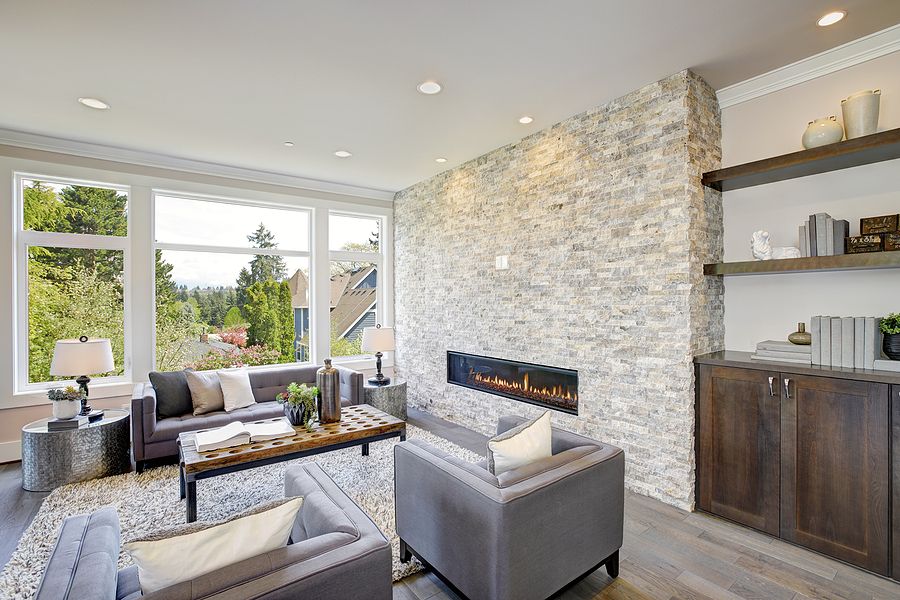 Newly renovated great room features a floor to ceiling stone fireplace gray tufted sofa paired with two gray armchairs over fluffy rug.