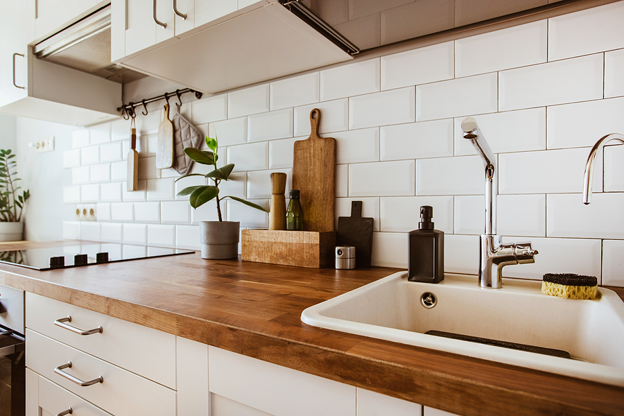 Kitchen brass utensils, chef accessories. Hanging kitchen with white tile backsplash and wood tabletop. Small green plant on kitchen counter.